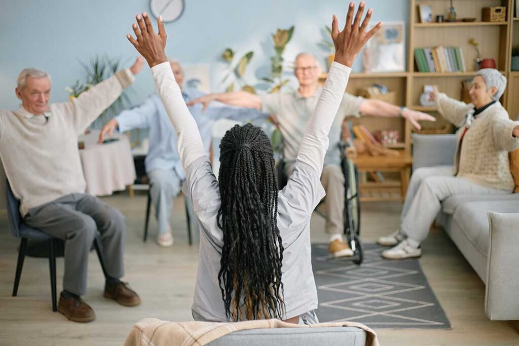 Seniors participating in a gentle chair yoga class with an instructor guiding movement in a brightly lit common room.